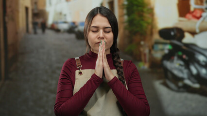 Waitress praying with eyes closed in an outdoor street setting wearing apron peaceful thoughtful...