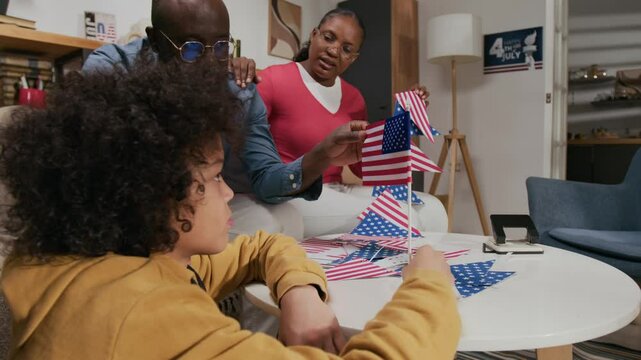 Zoom in view of gen alpha kid listening to dad telling him about history of American flag when they holding it sitting around white table on independence day, wife holding hand on husbands shoulder - Powered by Adobe