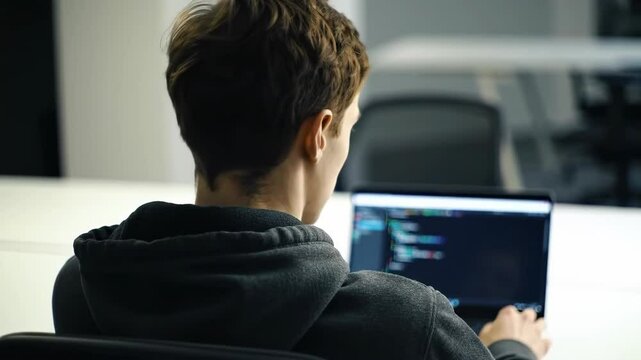 Back view of a young developer working on a laptop in a modern office He is coding or programming with a focused expression creating software in a bright workspace wearing a hoodie
