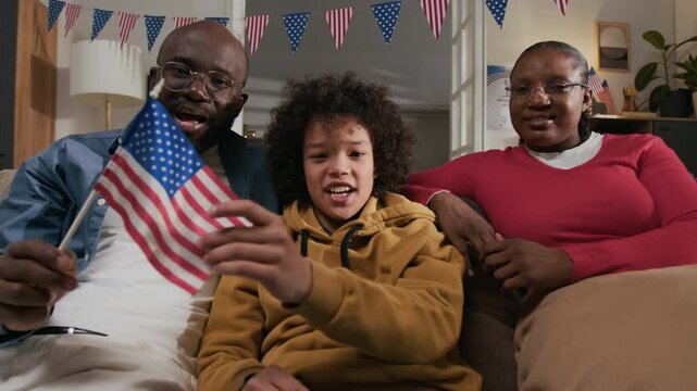 POV of cheerful Black family recording video message congratulating relatives on independence day in living room decorated with posters, patriotic garland and flags