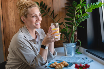Woman enjoying a fresh glass of orange juice at a kitchen table with croissants and strawberries,...