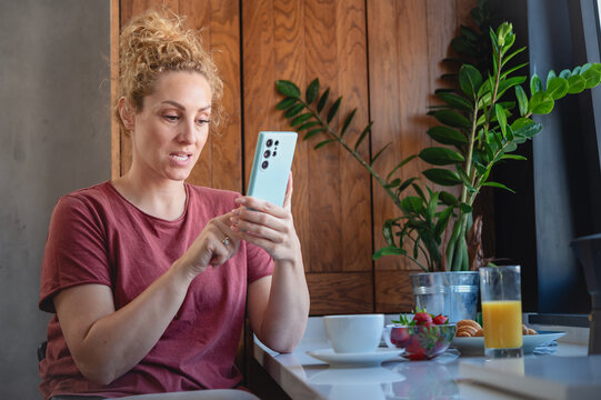 Fototapeta Nervous woman with curly hair looks at smartphone in modern kitchen, sitting by a breakfast table with juice, coffee, and fruit, appearing anxious or uncertain about a message.