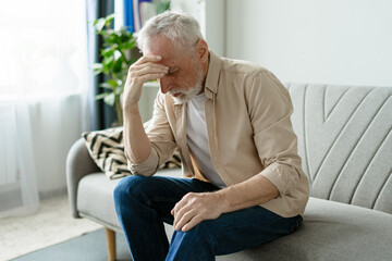 Worried senior man sitting on sofa at home touching his forehead