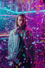 Man in silver glitter jacket standing under confetti at nightclub with neon lighting. Studio fashion portrait with LGBT expression theme. Design for poster, greeting card, postcard, banner. Front view