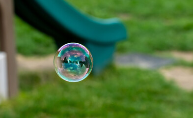 Single soap bubble with iridescent rainbow colors reflecting garden landscape floating in mid-air against blurred green yard background. Concept of summer childhood memories.