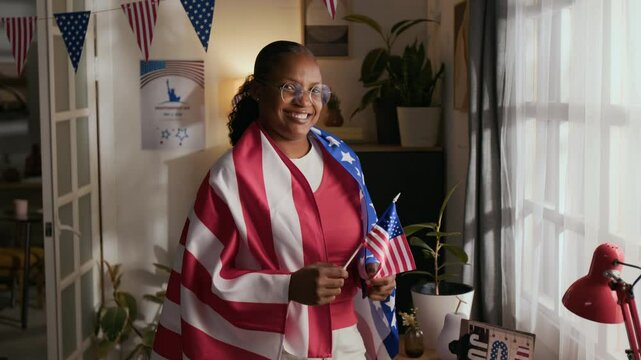 Portrait of joyful Black millennial woman wearing round glasses smiling for camera with American flags on Fourth of July at home
