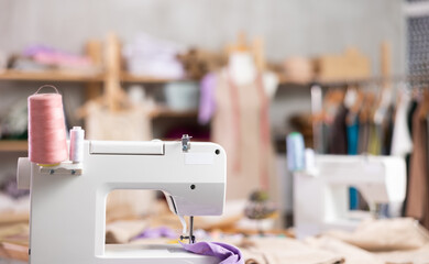 Closeup of modern sewing machine with pink and white thread spools in tailoring studio, with unfinished dress on mannequin, fabric shelves, and garment rack on softly blurred background..