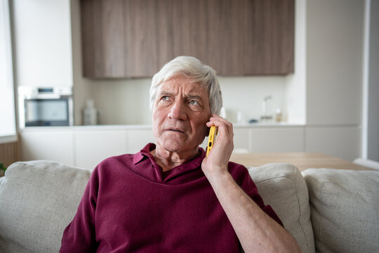 Distrustful and anxious senior man frowns during emergency call, listening to pressured demands for banking information, sitting at sofa. Fraud warning, mislead, personal data risk, financial scam