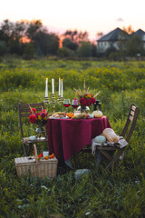 Beautiful decorated candlelight fall dinner, covered with a red tablecloth with flowers, pumpkins, candles, wine glasses. Outdoor in field near the forest in nature. Wedding banquet, sunset, evening