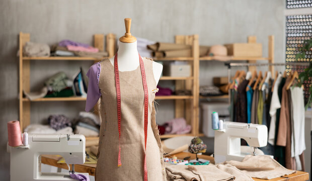 Mannequin with pinned unfinished linen dress and measuring tape in sewing studio, surrounded by fabric shelves, garment rack, and sewing tools, capturing creative tailoring atmosphere.. - Powered by Adobe