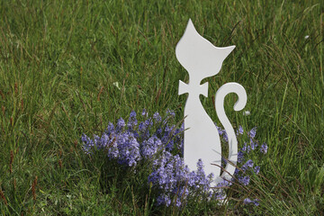 A wooden silhouette of a white cat sitting in the grass among purple flowers.
