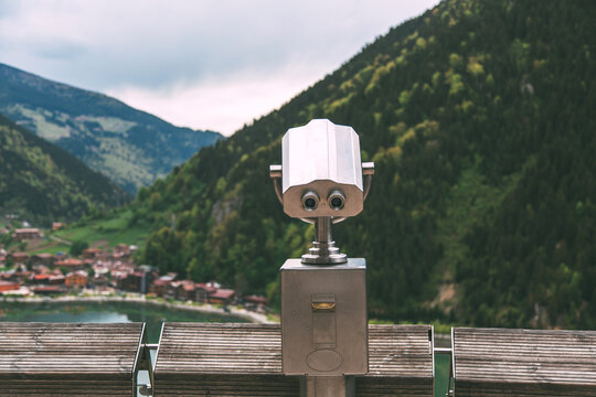 A coin-operated binocular viewer overlooks a scenic mountain valley and lake, offering panoramic views of a picturesque village surrounded by lush green hills.