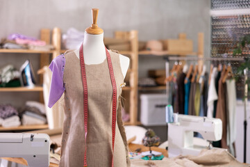 Mannequin with pinned unfinished linen dress and measuring tape in sewing studio, surrounded by fabric shelves, garment rack, and sewing tools, capturing creative tailoring atmosphere..