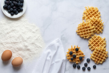 View from above of a white marble slab with ingredients for homemade waffles. In addition to flour and eggs, blueberries and ready-made waffles are sifted. There is space for text in the middle.