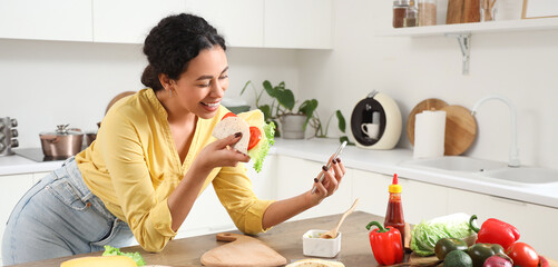 Young African-American woman with mobile phone eating tasty taco in kitchen