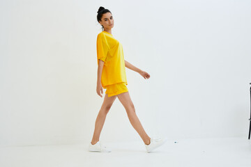 Young woman walking confidently indoors in bright yellow casual outfit with white sneakers against a minimalist white background, showcasing modern fashion style.