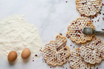 View from above of a white marble slab with ingredients for homemade waffles. Ready-made waffles can be seen next to flour and eggs. The pastries are decorated with dried flower petals.