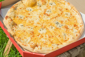 Pizza with cheese in a cardboard box on a bench on the grass. Picnic, food delivery.