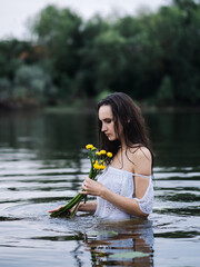 Beautiful young woman in white dress posing in river. Holding bouquet of water lilies. Brunette with long hair