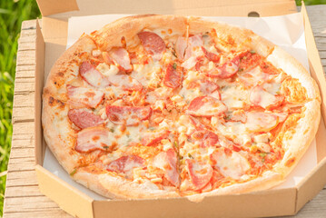 Meat pizza with cheese in a paper box on a wooden board on the grass.