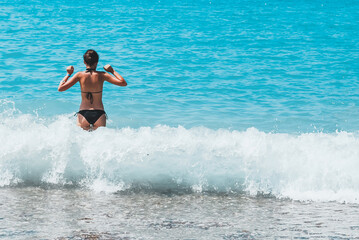 Young woman in black bikini playing in turquoise sea waves on a sunny beach, rear view, summer freedom and vacation mood concept, copyspace