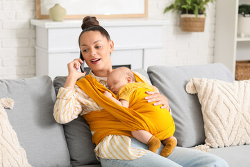 Young mother with cute little baby in sling talking by mobile phone at home