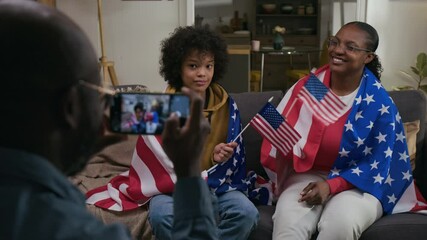Over the shoulder view of bald man taking horizontal smartphone picture of son and wife waving small US flags and smiling at camera on couch in living room - Powered by Adobe