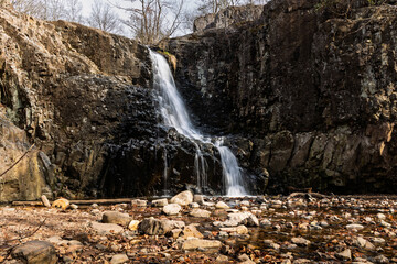 Hemlock Falls in New Jersey cascading over dark rocks into a shallow pool. A wide view of the waterfall flowing through rugged cliffs into a rocky stream bed in a natural wooded area.