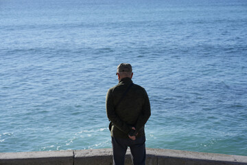 Unrecognizable senior man standing alone on embankment, watching the sea from behind. His posture...