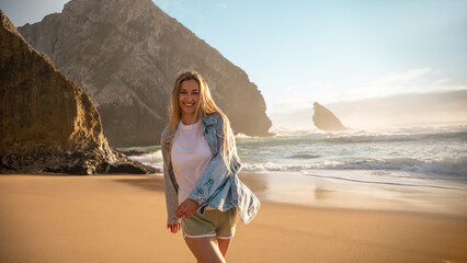Woman in denim shirt walking on sandy beach near large rock formations. Smiling lady with long blond hair looking at camera with waves in background. Warm sunlight casting soft glow on scene