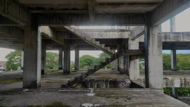 An abandoned concrete building with exposed beams and a worn, unfinished staircase. Natural light filters through, revealing distant green trees beyond the open structure.