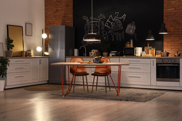 Interior of stylish kitchen with counters, dining table and glowing lamps at night