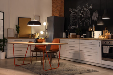 Interior of stylish kitchen with counters, dining table and glowing lamps at night