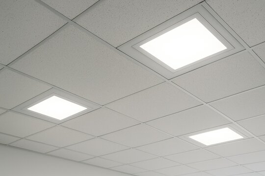 Detailed shot of sleek square recessed LED downlights seamlessly integrated into a modern office ceiling with acoustic white square tiles creating a clean and efficient lighting design.