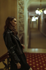Young woman in a black leather jacket stands thoughtfully in a vintage hallway with soft lighting. Stylish, moody portrait with cinematic atmosphere.