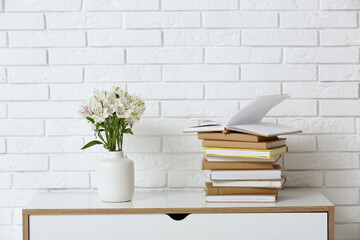 Stack of different books and vase with flowers on chest of drawers near light brick wall