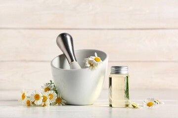 Bottle of chamomile cosmetic oil and mortar with flowers on white wooden background