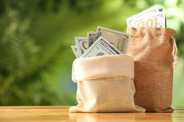 Fototapeta na wymiar Money bags with dollar banknotes on wooden table against blurred green background, closeup. Space for text