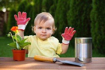 young child wearing gardening gloves sitting at wooden table with potted flower, gardening trowel, and empty tin can outdoors in garden