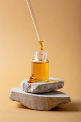 Glass bottle filled with golden oil with a dropper dispensing oil drops, placed on stacked natural stone slabs against a warm beige background