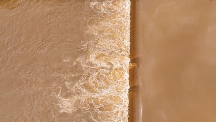 Drone view of brown floodwaters overflowing a barrier, demonstrating the widespread impact of heavy rains and flooding on natural water systems and surrounding habitats.
