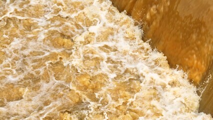 Aerial shot reveals brown water rushing down a waterfall or dam, highlighting the dynamic movement and natural processes of sediment-rich water in river ecosystems.
