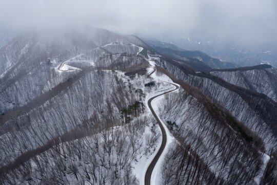 A winding mountain road covered with patches of snow surrounded by leafless trees and foggy mountains under a cloudy sky