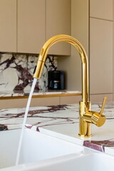modern kitchen sink with shiny gold faucet running water on marble countertop with beige cabinets in background