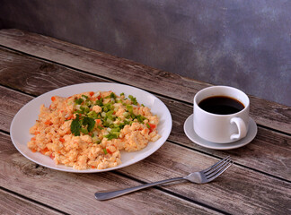 A plate of egg omelette fried with tomatoes and garnished with a sprig of parsley on a wooden table, next to it a fork and a cup of hot black coffee.
