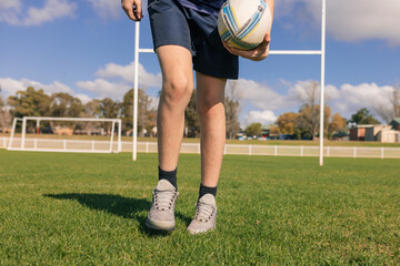 Waist-down view of junior rugby union player holding ball on field in front of goal posts