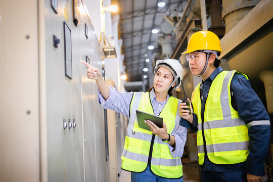 Two engineers in safety helmets and vests inspect electric switchgear panel in industrial field using tablet and walkie talkie near insulated gas equipment in grid environment