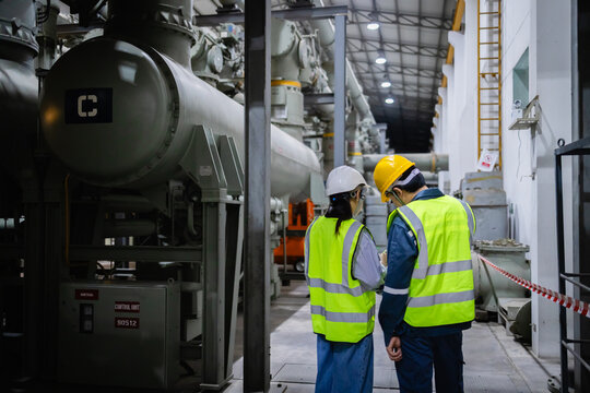 Two engineers wearing insulated safety helmets and reflective vests inspect electric grid switchgear in large industrial field with gas equipment and GIS components