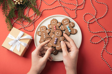 Hands holding gingerbread cookies on a white plate decorated with colorful sprinkles surrounded by holiday decor and gift on a red background. Top view