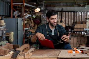 Male carpenter checking smartphone while holding clipboard in wood workshop, multitasking with...